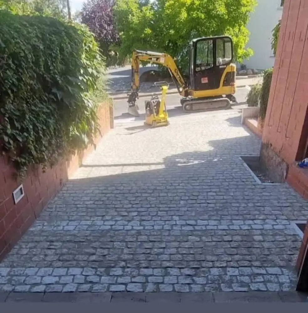 Neu verlegter Pflasterweg aus Natursteinpflaster zwischen Hauswand und Hecke, im Hintergrund ein gelber Minibagger von Christian Hopert Galabau
